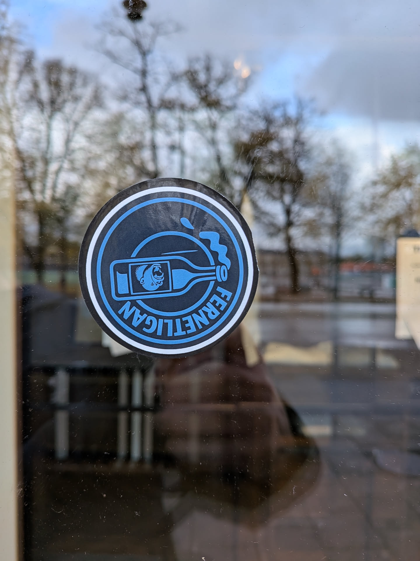 Street sticker The sticker is circular with a black outer ring and a white inner ring. Inside, the background is black. A blue outline depicts a bottle with a monkey's head visible inside. Blue liquid is splashing out of the bottle's neck. The text 'FERNETLIGAN' is written in blue, curved lettering around the bottom half of the sticker.