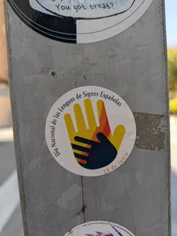 Street sticker The sticker is round and placed on a metallic surface. It shows a large yellow and orange hand with a smaller dark hand inside. The text 'Día Nacional de las Lenguas de Signos Españolas' (National Day of Spanish Sign Languages) circles the top of the design, and '14 de junio' (June 14th) is written at the bottom.
