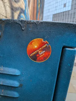 Street sticker A circular sticker featuring a dynamic gradient background that transitions from a bright, almost glowing orange at the top, suggesting a sun or sunrise, through fiery reds, to a dark, almost black hue at the bottom. The text "HORIZON RADIO" is prominently displayed in an arc across the upper half in white, with a subtle yellow outline. Below this, a classic red YouTube play button logo is visible, followed by the word "YouTube" in white and "24-7" in orange. The sticker is adhered to a textured, dark blue surface and shows signs of wear, including a small tear on its right edge and some minor scuffs.