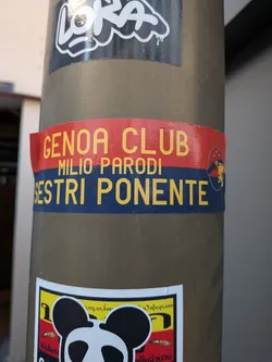 A rectangular sticker divided horizontally into red and blue bands. It features yellow text and a small circular crest on the right side.