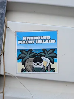 Street sticker A square sticker featuring a cartoon character in a black and white balaclava on a tropical beach. The character is flanked by a vintage leather football and a foaming beer mug, with palm trees and the ocean behind them. The German text 'HANNOVER MACHT URLAUB' is written in bold white letters across the blue sky.