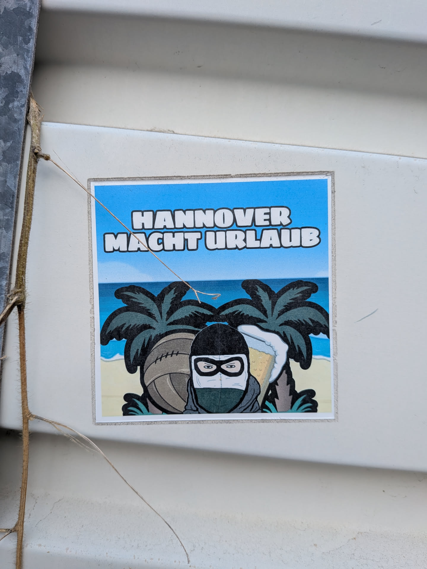 A square sticker featuring a cartoon character in a black and white balaclava on a tropical beach. The character is flanked by a vintage leather football and a foaming beer mug, with palm trees and the ocean behind them. The German text 'HANNOVER MACHT URLAUB' is written in bold white letters across the blue sky.