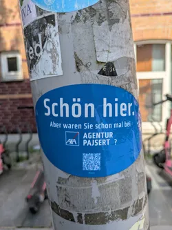 Street sticker A blue oval-shaped sticker is affixed to a weathered utility pole, which is also covered with remnants of other stickers. The sticker's design is clean and features white text on a solid blue background. The main text, in German, reads "Schön hier. Aber waren Sie schon mal bei- AXA AGENTUR PAJSERT ?" which translates to "It's nice here. But have you ever been to- AXA AGENCY PAJSERT ?". Below the agency name, there is a clear QR code with the small text "vCard" underneath it, indicating it's for contact information. The pole itself shows signs of age and previous adhesive marks, typical of urban environments.