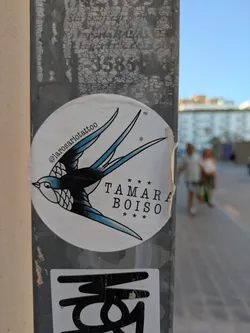 Street sticker This round sticker, affixed to a metal pole, showcases a classic tattoo-style swallow. The bird is rendered in black and blue, with detailed shading on its wings and chest. The text "TAMARA BOISO" is printed in a simple font, surrounded by small stars. Above the bird, the Instagram handle @larosariotattoo is visible, indicating the artist or tattoo parlor responsible for the design.