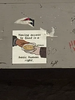 Street sticker The sticker is square-shaped with a white background. It features a simple, line-drawn illustration of two hands. One hand, with a brown skin tone, is offering a bowl of soup to another hand, which is receiving it. The text above the illustration reads 'Having access to food is a' in black, sans-serif font. Below the illustration, the text continues 'basic human right.' in the same font. To the right of the illustration, a social media handle '@fnbwro' is visible in smaller text. The sticker appears to be slightly creased and adhered to a textured, grey surface, with some other peeling stickers or marks around it.