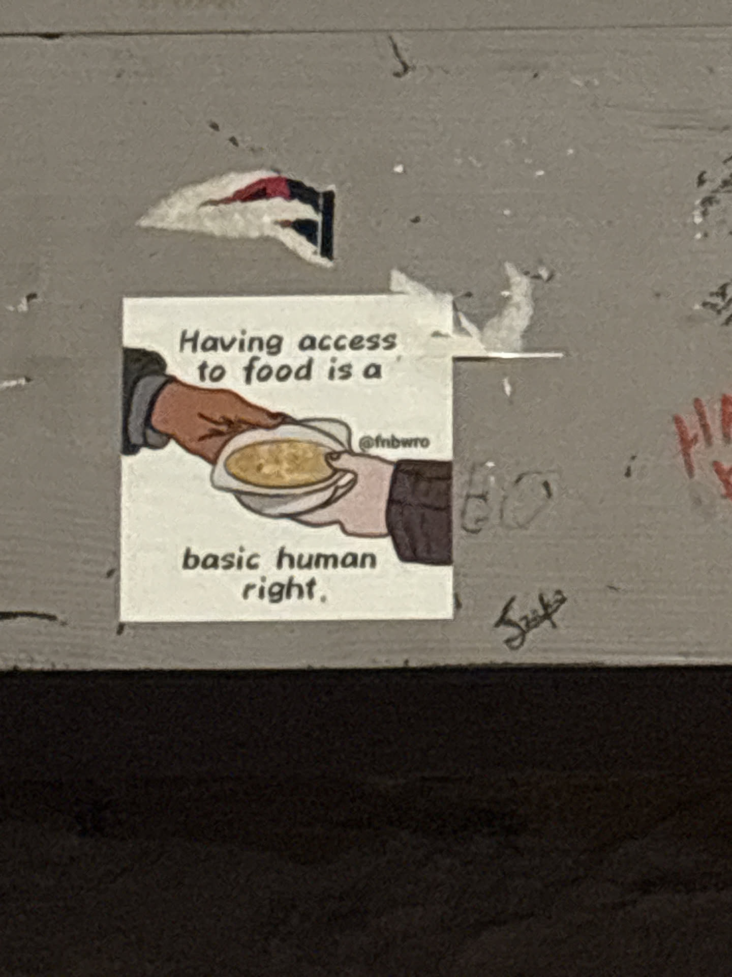 Street sticker The sticker is square-shaped with a white background. It features a simple, line-drawn illustration of two hands. One hand, with a brown skin tone, is offering a bowl of soup to another hand, which is receiving it. The text above the illustration reads 'Having access to food is a' in black, sans-serif font. Below the illustration, the text continues 'basic human right.' in the same font. To the right of the illustration, a social media handle '@fnbwro' is visible in smaller text. The sticker appears to be slightly creased and adhered to a textured, grey surface, with some other peeling stickers or marks around it.