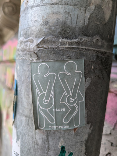 A humorous sticker on a weathered pole features stick figures holding toilet paper rolls over their rear ends. The text reads 'store restroom'.