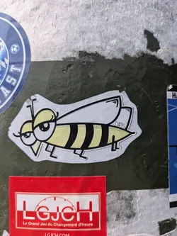 Street sticker A charming cartoon sticker of a bee or wasp, featuring a yellow and black striped body, large, expressive eyes with heavy eyelids giving it a 'sassy' or 'bored' look, and delicate, transparent wings. The artist's name 'DIRK' is subtly written on one of its wings. The sticker is affixed to a textured, dark green surface, with fragments of other stickers visible around it.