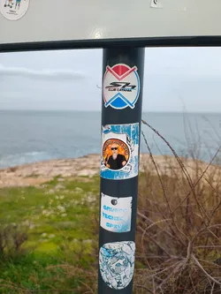 Street sticker A circular sticker featuring a cartoon illustration of a man with a long, flowing white beard and dark sunglasses. He wears a black t-shirt and holds a brown beer bottle in his right hand. The character is depicted against a vibrant orange and yellow gradient background, encircled by a white border. The text 'CERTIFICADO POLLA VIEJA' is written in black capital letters along the top curve of the white border. The sticker shows signs of wear, with some blue and white remnants of another sticker underneath or around it, suggesting it's adhered to a textured surface.