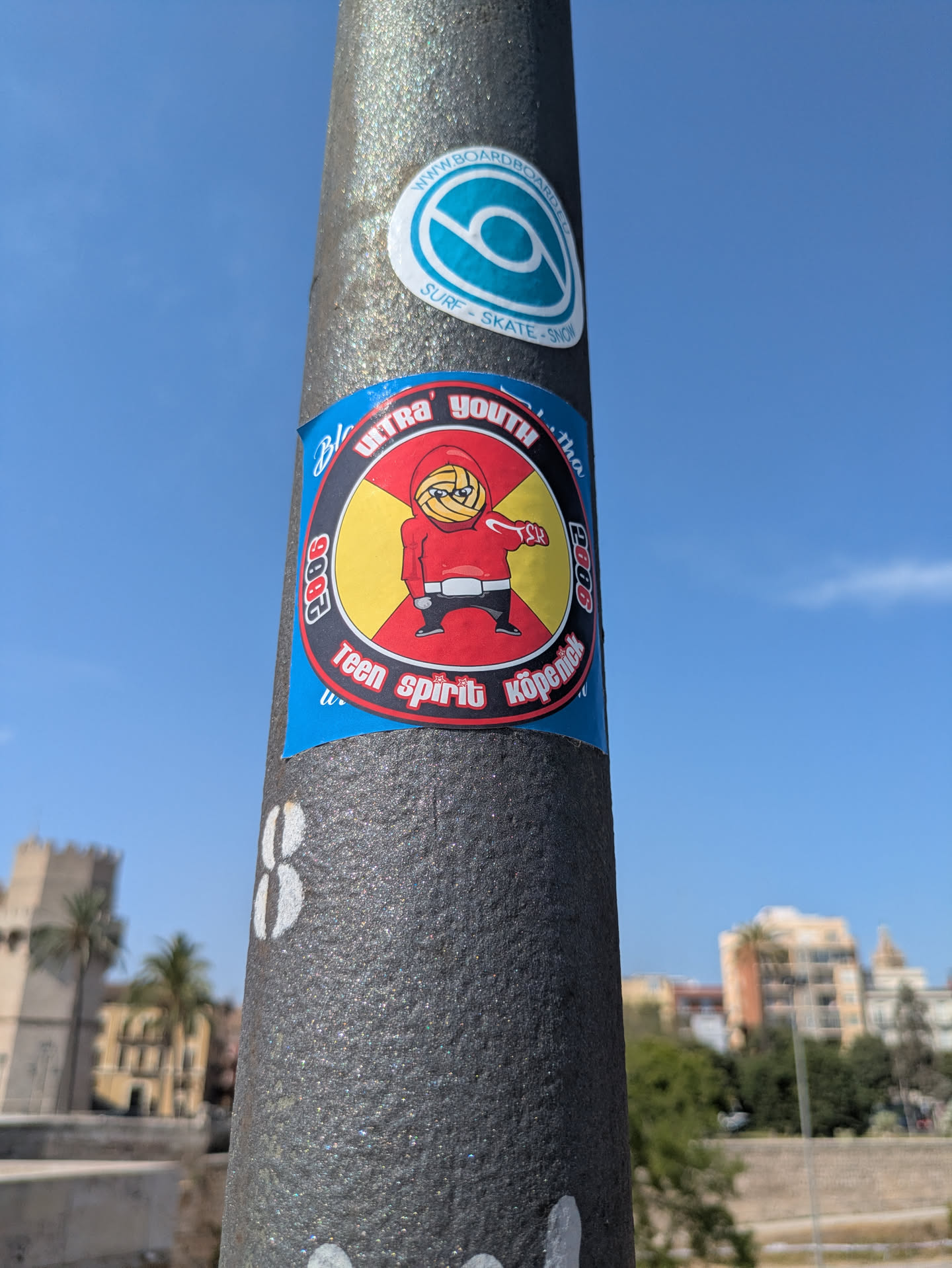 This sticker features a character with a volleyball for a face, wearing a red hoodie and black pants. The sticker is red and yellow, with the words "Ultra Youth," "Teen spirit Köpenick," and "2006" written around the character. The sticker is pasted on a metal pole. There is another round sticker above it with the text "WWW.BOARDBOARD.EU SURF-SKATE-SNOW"