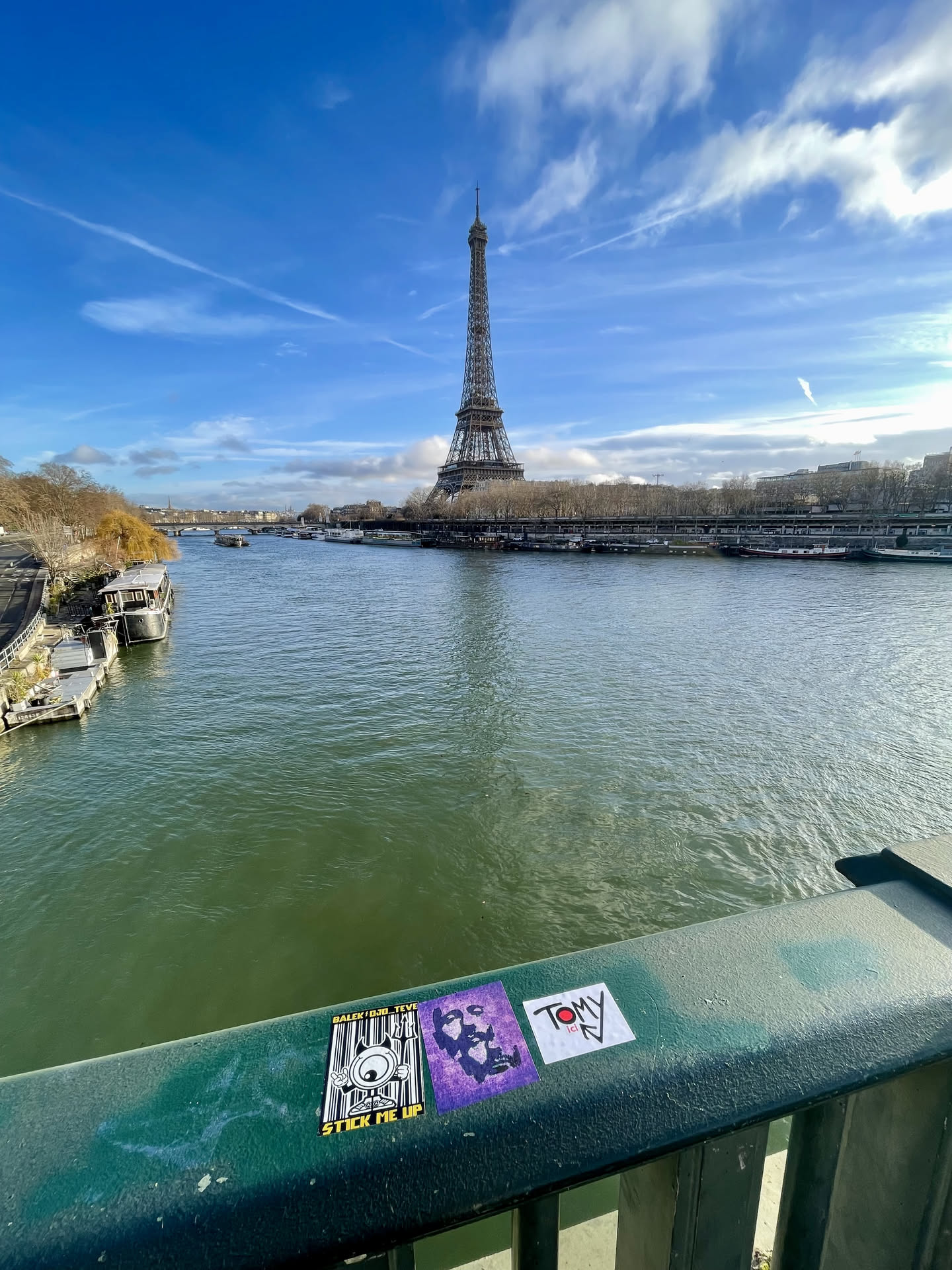Three distinct stickers are affixed to a weathered green metal bridge railing. The leftmost sticker is rectangular, featuring a black and white design with a yellow top border. It depicts a cartoonish, one-eyed monster character with raised hands, set against a barcode pattern background. Text at the top reads 'BALEK | OJO_TEVE' and 'STICK ME UP' at the bottom. The middle sticker is square and predominantly purple, showcasing an abstract, dark-colored face or figure in a rough, possibly stencil-like style. The rightmost sticker is a white square with prominent black text 'TOMY' in capital letters, a red dot above the 'O', and a small downward arrow pointing to 'ici' below the name.