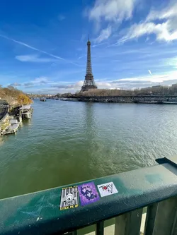 Street sticker Three distinct stickers are affixed to a weathered green metal bridge railing. The leftmost sticker is rectangular, featuring a black and white design with a yellow top border. It depicts a cartoonish, one-eyed monster character with raised hands, set against a barcode pattern background. Text at the top reads 'BALEK | OJO_TEVE' and 'STICK ME UP' at the bottom. The middle sticker is square and predominantly purple, showcasing an abstract, dark-colored face or figure in a rough, possibly stencil-like style. The rightmost sticker is a white square with prominent black text 'TOMY' in capital letters, a red dot above the 'O', and a small downward arrow pointing to 'ici' below the name.