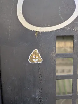 A poop emoji sticker is stuck on a black metal surface. The poop emoji is cartoonish, with big blue eyes and two buck teeth.