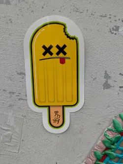 Street sticker This vibrant sticker portrays a yellow popsicle with a distinct personality. It has a cartoonish face featuring black 'X's for eyes and a small red tongue peeking out from a thin black line that serves as a mouth, giving it a playful yet 'dead' or 'finished' appearance. A bite mark is visible on the upper right side of the popsicle. The light brown popsicle stick has the word "BESTe" written vertically in black, likely serving as an artist's signature or brand. The entire design is outlined in black and a bright lime green, and is placed on a light grey, textured wall.