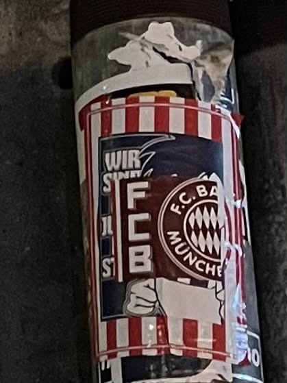 The sticker features a prominent design dominated by the colors red, white, and dark blue. The background consists of alternating red and white vertical stripes. Centrally, the official circular logo of FC Bayern München is displayed, with 'F.C. BAYERN MÜNCHEN' written around its perimeter and the traditional blue and white diamond pattern in the center. To the left of the main logo, vertical text reads 'WIR SIND' at the top, followed by 'ULTRAS' (partially obscured but discernible), and then 'FCB' stacked vertically. Below the 'FCB' text, a stylized white fist is depicted. The sticker shows signs of wear, including peeling edges and creases, indicating it is a physical sticker applied to a cylindrical object.