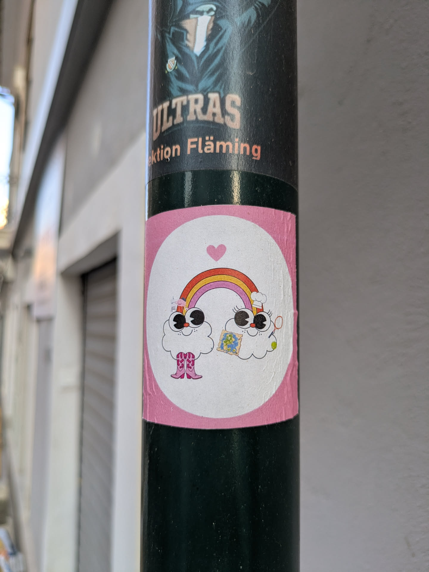 This is a round sticker on a lamppost. It features two cloud characters, each with large eyes and smiling faces. A colorful rainbow arcs between them, topped with a small pink heart. The cloud on the left is wearing pink cowboy boots, and the one on the right holds a small map.