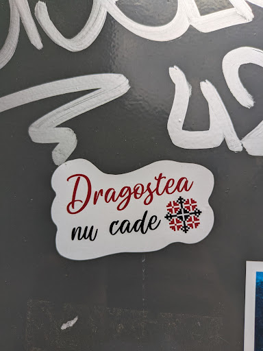 A white sticker with rounded, irregular edges. The text "Dragostea nu cade" is printed on the sticker in a mix of red and black fonts. A traditional Romanian design element, resembling a geometric flower, is included as part of the design.