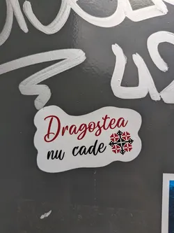 Street sticker A white sticker with rounded, irregular edges. The text "Dragostea nu cade" is printed on the sticker in a mix of red and black fonts. A traditional Romanian design element, resembling a geometric flower, is included as part of the design.