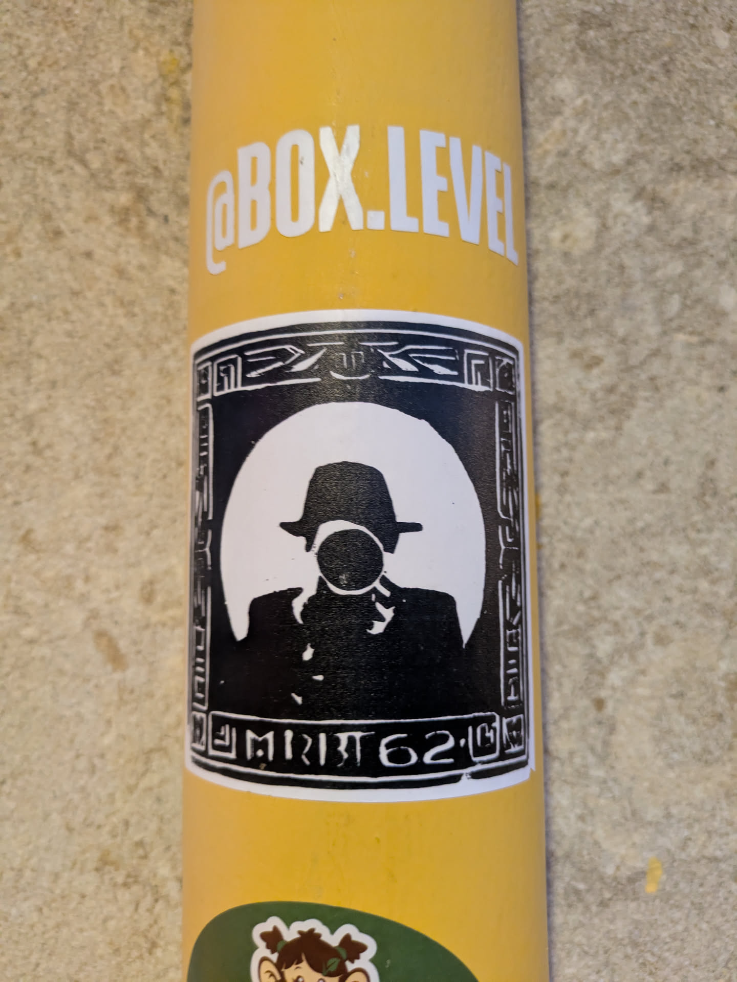 The sticker features a black and white silhouette of a man with a hat blowing a bubble, framed in a black border with designs that resemble ancient Egyptian hieroglyphs. The background is a white circle. There is another sticker on the bottom of the yellow surface.