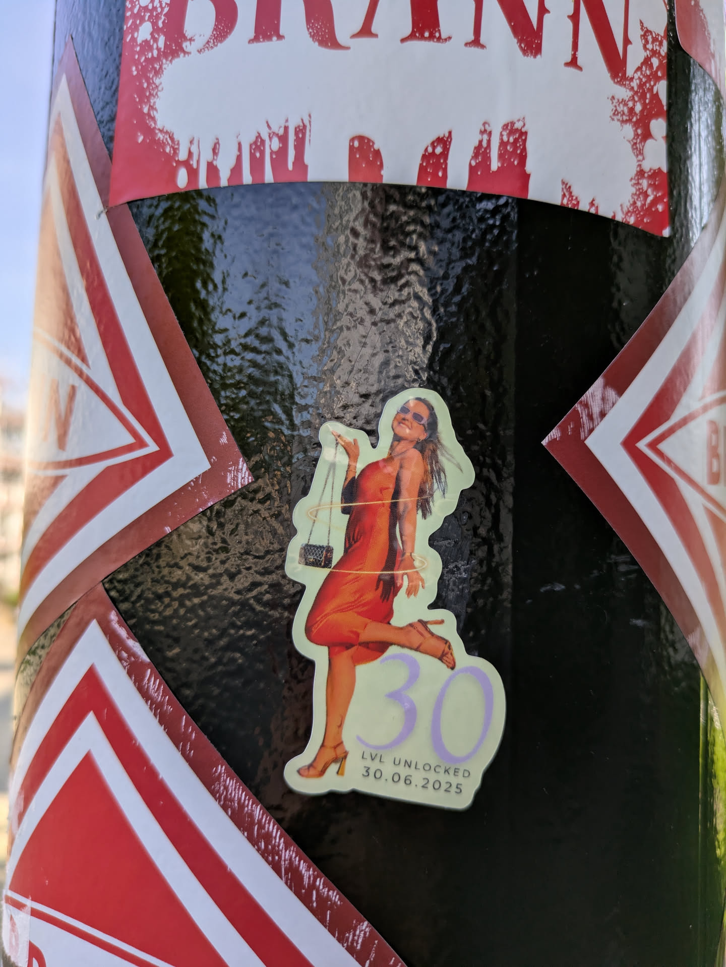 The sticker features a cheerful woman in a flowing orange dress and sunglasses, posing dynamically with a small black purse. A subtle light green aura or ring encircles her. Below the figure, the large number "30" is prominently displayed in light purple, accompanied by the text "LVL UNLOCKED" and the date "30.06.2025", indicating a future 30th birthday celebration with a gaming achievement motif. The sticker has a light green outline and is placed on a textured black surface.
