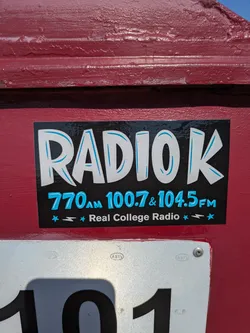 The sticker is rectangular with a black background that appears to have a slightly distressed or textured finish. The main text 'RADIO K' is displayed prominently in large, bold white letters with light blue outlines, giving them a playful, slightly bubbly appearance. Below the main logo, in smaller light blue text, are the frequencies '770AM 100.7 & 104.5FM'. At the bottom, in white text, it reads 'Real College Radio', flanked by small light blue stars and lightning bolt symbols. The sticker is affixed to a textured, dark red painted surface.