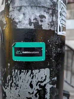 Street sticker The sticker is a rectangular shape with slightly rounded corners, featuring a bright, almost neon green background. A prominent horizontal black bar runs across the center. On the left side of this black bar, there is a minimalist logo composed of two outlined green triangles, resembling mountains. To the right of the logo, the word "makinlocations" is printed in white, sans-serif text. The sticker shows minor signs of wear, particularly at the top left corner where it is slightly peeled.