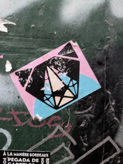 This sticker features a geometric diamond design. The diamond has a white center, black outline, and blue facets, set against a pink square background. The sticker is placed on a dark green wall with signs of wear and tear.
