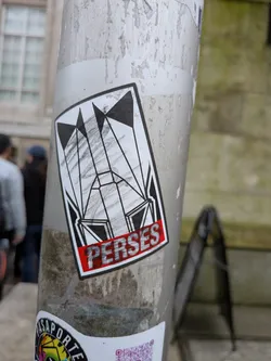 Street sticker The sticker is a rectangular design with slightly rounded corners, featuring a bold black outline. The main graphic is set against a white background, composed of intricate black geometric shapes that resemble a stylized, abstract structure or a series of flags/banners. These shapes create a sense of depth and angularity. Within some of the white spaces, there are subtle grey, pencil-like hatch marks, adding texture. At the bottom, a vibrant red banner with a black outline contains the word 'PERSES' in white, blocky, textured letters, also outlined in black.