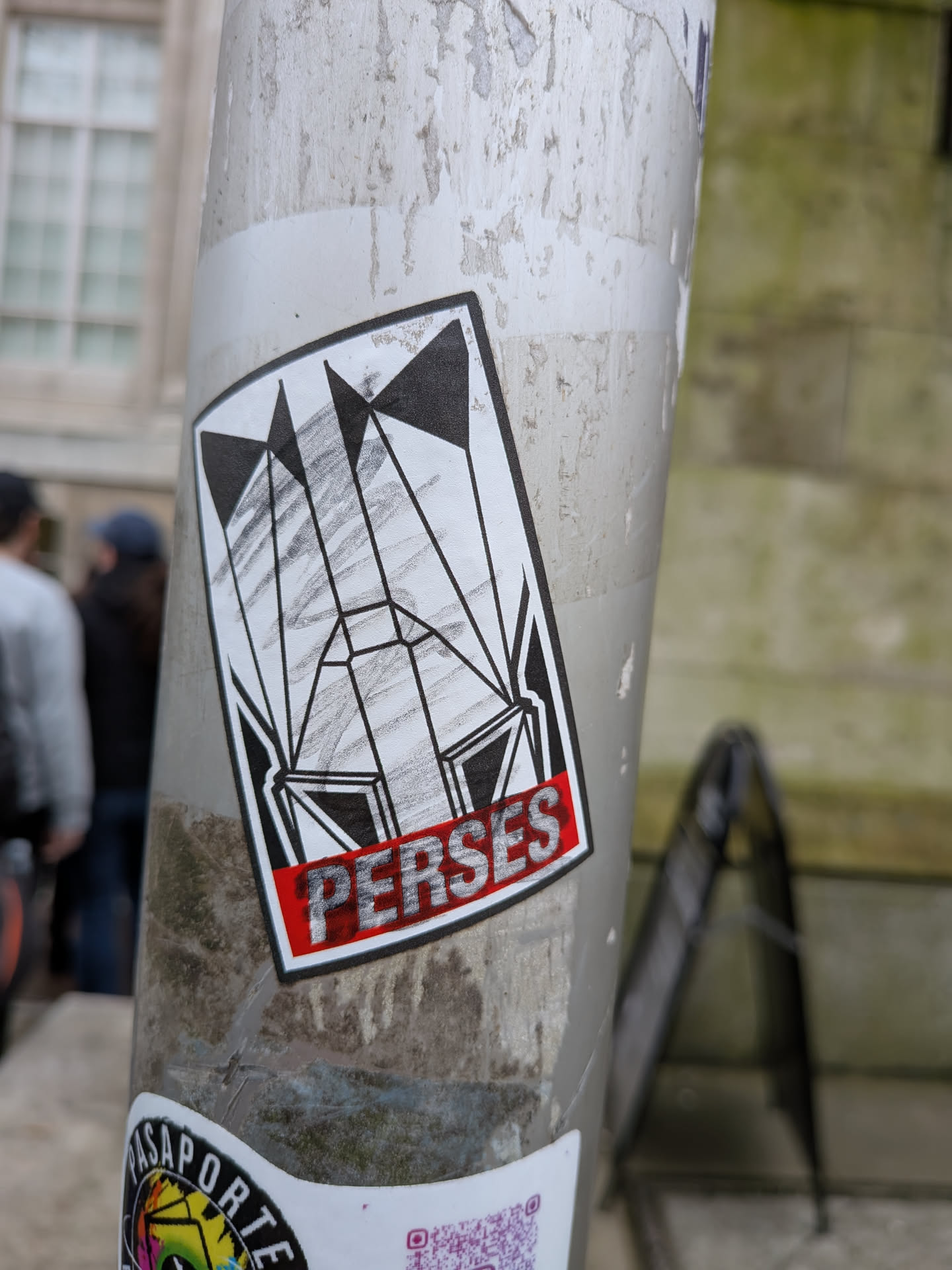 The sticker is a rectangular design with slightly rounded corners, featuring a bold black outline. The main graphic is set against a white background, composed of intricate black geometric shapes that resemble a stylized, abstract structure or a series of flags/banners. These shapes create a sense of depth and angularity. Within some of the white spaces, there are subtle grey, pencil-like hatch marks, adding texture. At the bottom, a vibrant red banner with a black outline contains the word 'PERSES' in white, blocky, textured letters, also outlined in black.