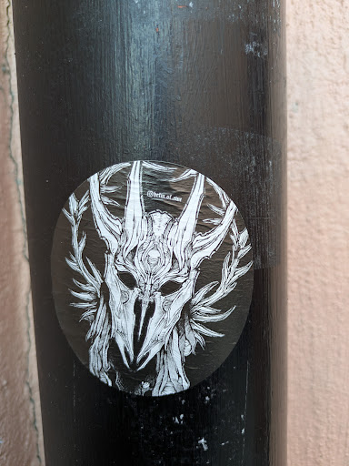 Street sticker This round sticker showcases a detailed illustration of a skull mask or helmet, reminiscent of ceremonial attire. The mask is surrounded by antler-like structures and foliage, creating an organic yet eerie aesthetic. The background is black, providing a stark contrast to the white linework. It's located on what appears to be a black metal or painted pole.