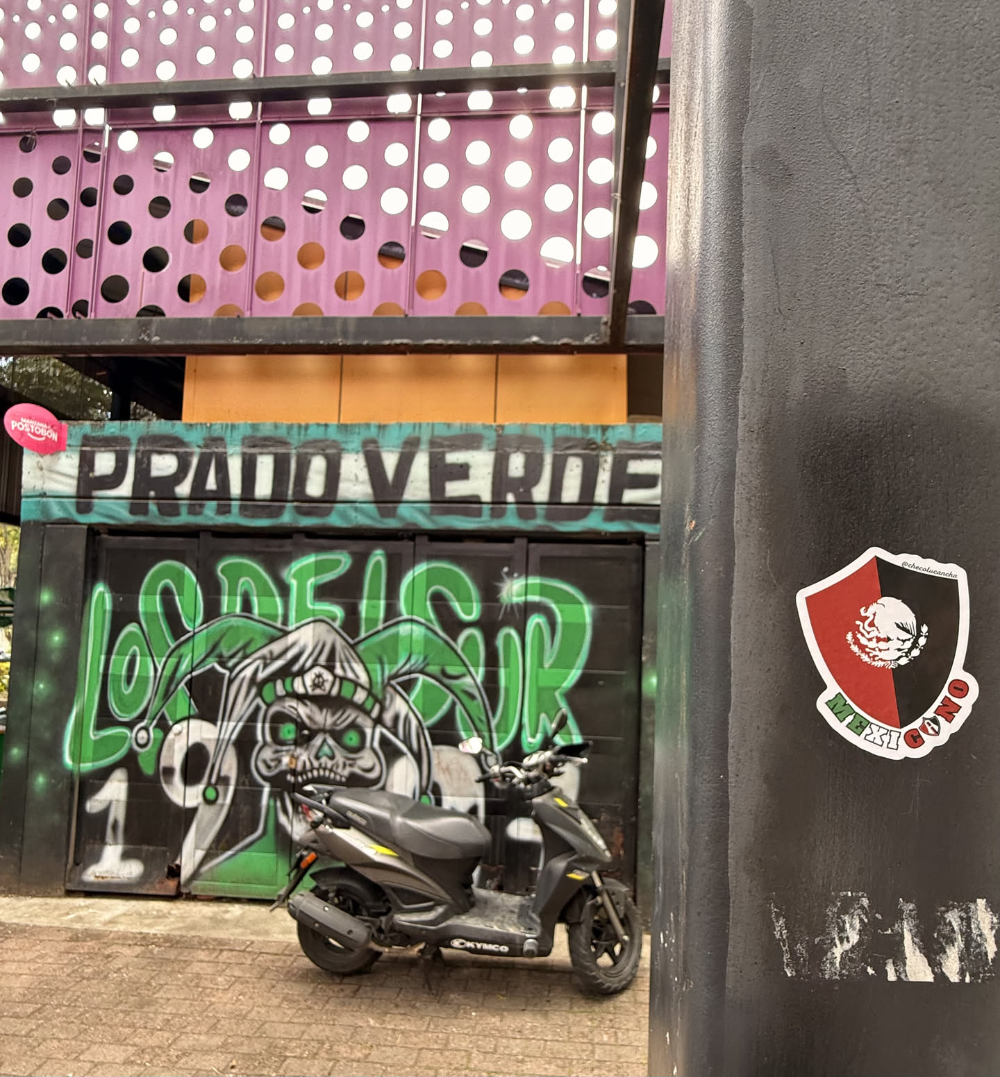 The sticker is shaped like a shield, divided diagonally into red and black halves. The top left portion is red, and the bottom right is black. In the center, a white depiction of the Mexican national emblem (an eagle devouring a snake, perched on a nopal cactus) is prominently displayed. Along the bottom edge, the word "MEXI" is written in white on the red half, and "GONO" is written in white on the black half, forming "MEXIGONO". A thin white border outlines the entire sticker. At the very top, in tiny white text, is a social media handle.