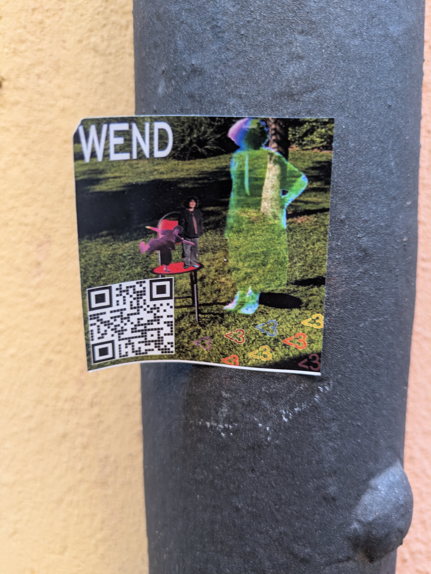 The sticker shows a grassy outdoor scene. On the left, a person stands on a small, red, table-like chair. Beside them is another figure, translucent and grassy-green. The text 'WEND' is visible at the top left. The bottom of the sticker shows small colored hearts. A QR code is also present.