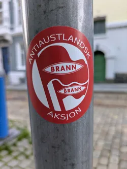 Street sticker A vibrant red circular sticker with white text and graphics is affixed to a grey metal pole. The sticker features two stylized white flag or banner shapes, each containing the word "BRANN" in red text. Arched above the central design is the white text "ANTIAUSTLANDSK", and below it, also in white, is "AKSJON". This sticker appears to be related to the SK Brann football club from Bergen, Norway, and possibly an 'Anti-Eastland' regional identity or rivalry action.
