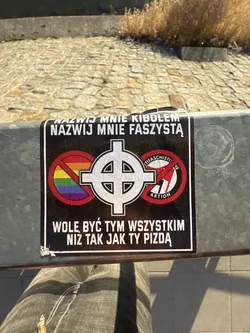 Street sticker A rectangular sticker with a black background and a thin white border. At the top, two lines of white uppercase text read 'NAZWIJ MNIE KIBOLEM' and 'NAZWIJ MNIE FASZYSTĄ'. The central part of the sticker features a large white Celtic cross. To the left of the cross, there is a red circle with a diagonal line crossing out a rainbow flag. To the right, another red circle with a diagonal line crosses out the Antifa symbol, which includes the text 'ANTIFASCHISTI' and 'AKTION'. Below these symbols, two more lines of white uppercase text read 'WOLE BYĆ TYM WSZYSTKIM' and 'NIZ TAK JAK TY PIZDĄ'. The sticker appears slightly worn and dusty, applied to a textured metal surface.