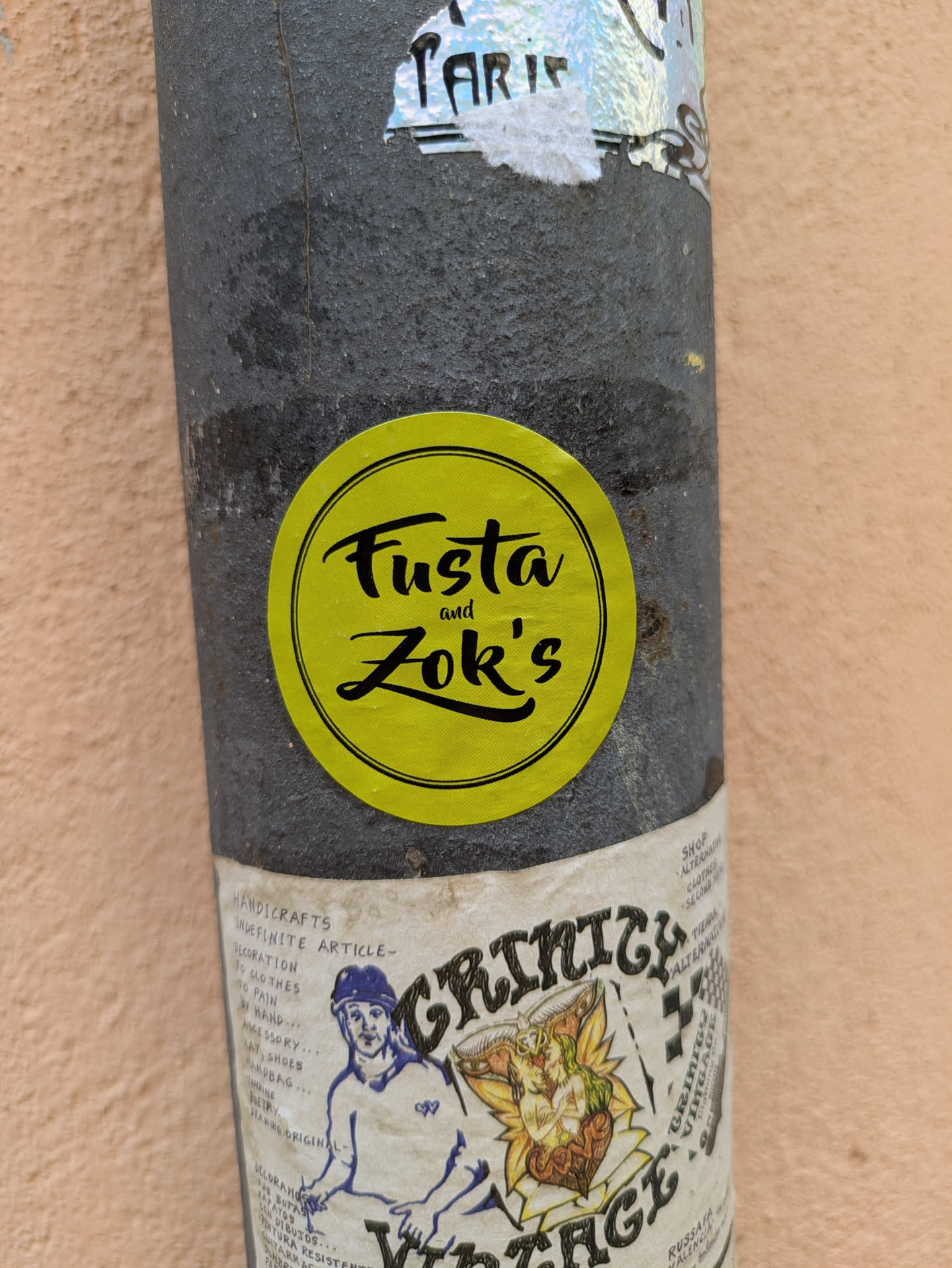 A round yellow sticker with the words "Fusta and Zok's" written in black script. It is stuck on a cylindrical pole along with other stickers, including one for "Grinicy Vintage".