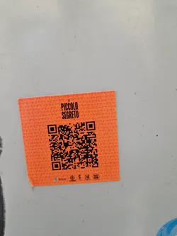 Street sticker Orange square sticker with a QR code in the center. The text "Un Piccolo Segreto" is printed on top of the QR code. There are some small logos at the bottom of the sticker.