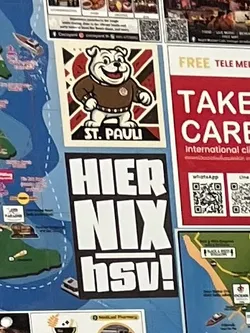 Street sticker The central sticker is composed of two vertically stacked rectangular parts. The top part features a smiling, cartoonish white bulldog mascot wearing a brown sweater with white trim and a small emblem on its chest. The bulldog stands against a background of radiating dark brown and red-brown stripes, all enclosed within a white border. Below the bulldog, on a black strip, is the white text 'ST. PAULI'. The bottom part of the sticker is a black rectangle with large, bold white text arranged vertically: 'HIER', 'NIX', and 'hsv!' with an exclamation mark, where 'hsv' is in lowercase and underlined.