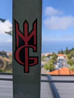 Street sticker A die-cut sticker featuring a red stylized 'MG' monogram with a black outline, affixed to a light green metal post.