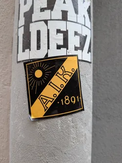 A square sticker featuring the AIK crest. It displays a diagonal yellow stripe containing the letters 'A.I.K.', a yellow sun in the upper-left corner, and the year '1891' in the lower-right corner, set against a black background.