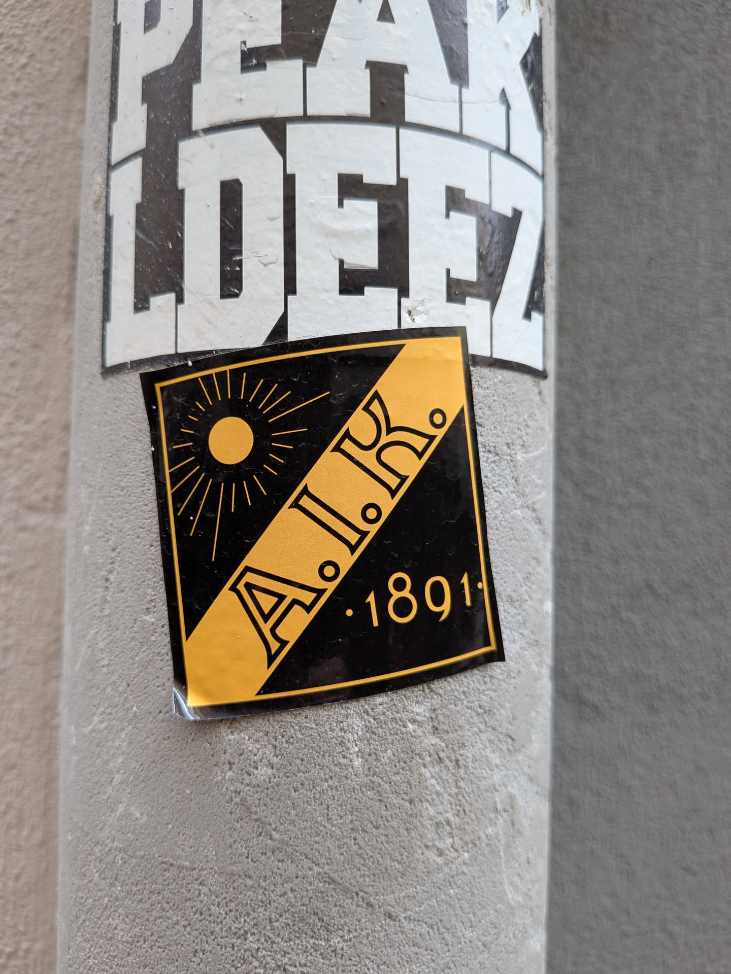 A square sticker featuring the AIK crest. It displays a diagonal yellow stripe containing the letters 'A.I.K.', a yellow sun in the upper-left corner, and the year '1891' in the lower-right corner, set against a black background.