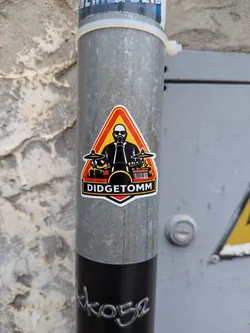 Street sticker Triangular sticker depicting a bald, sunglass-wearing musician playing a didgeridoo and drums. Features a red, orange, and yellow background with 'DIDGETOMM' in white text on a black banner at the bottom.