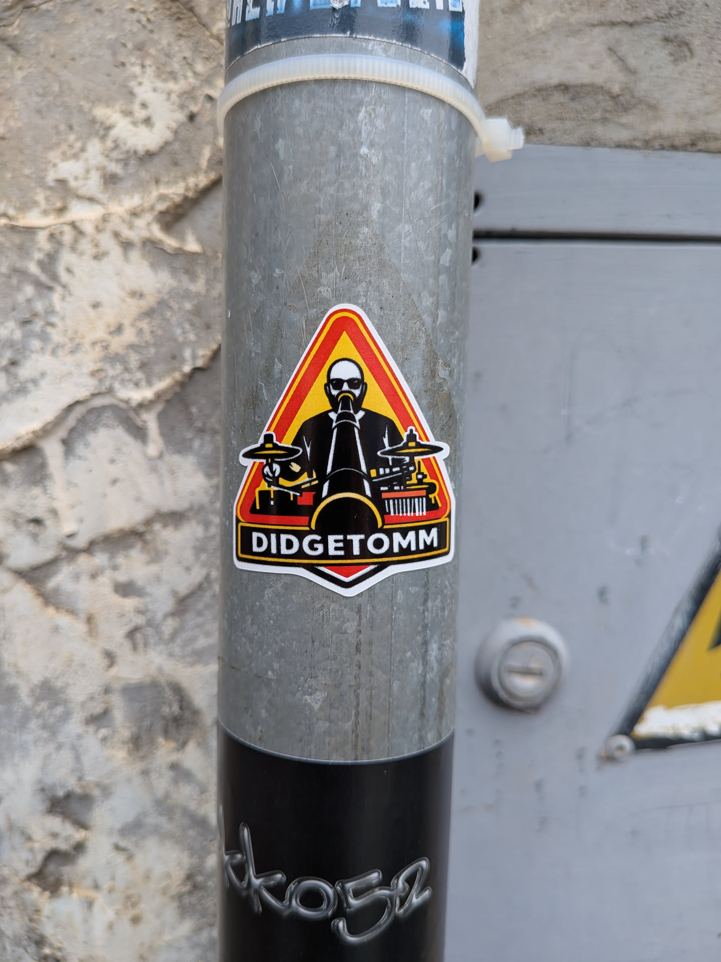 Triangular sticker depicting a bald, sunglass-wearing musician playing a didgeridoo and drums. Features a red, orange, and yellow background with 'DIDGETOMM' in white text on a black banner at the bottom.