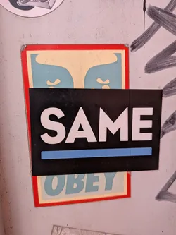 Street sticker A black rectangular sticker featuring the white text 'SAME' with a blue underline. It is layered on top of a vertical 'OBEY' sticker with a red border and blue-cream graphic.