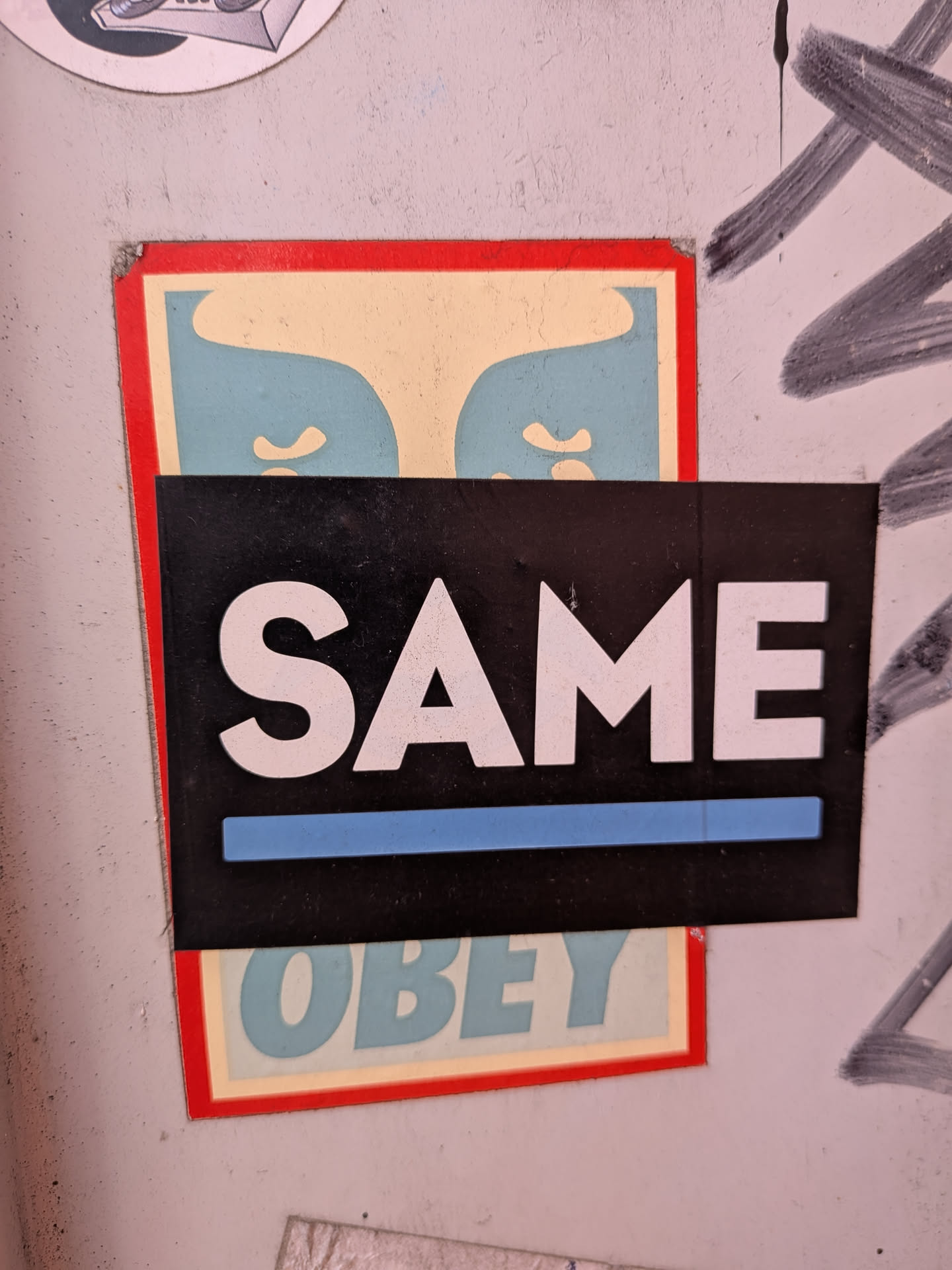 A black rectangular sticker featuring the white text 'SAME' with a blue underline. It is layered on top of a vertical 'OBEY' sticker with a red border and blue-cream graphic.