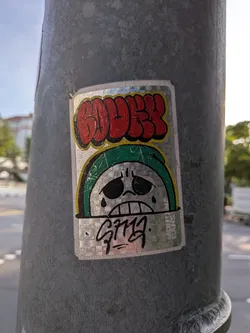 Street sticker A rectangular holographic sticker affixed to a metal pole. It features red bubble letters outlined in yellow at the top, a sad green-hooded character in the center, and a black marker tag at the bottom.