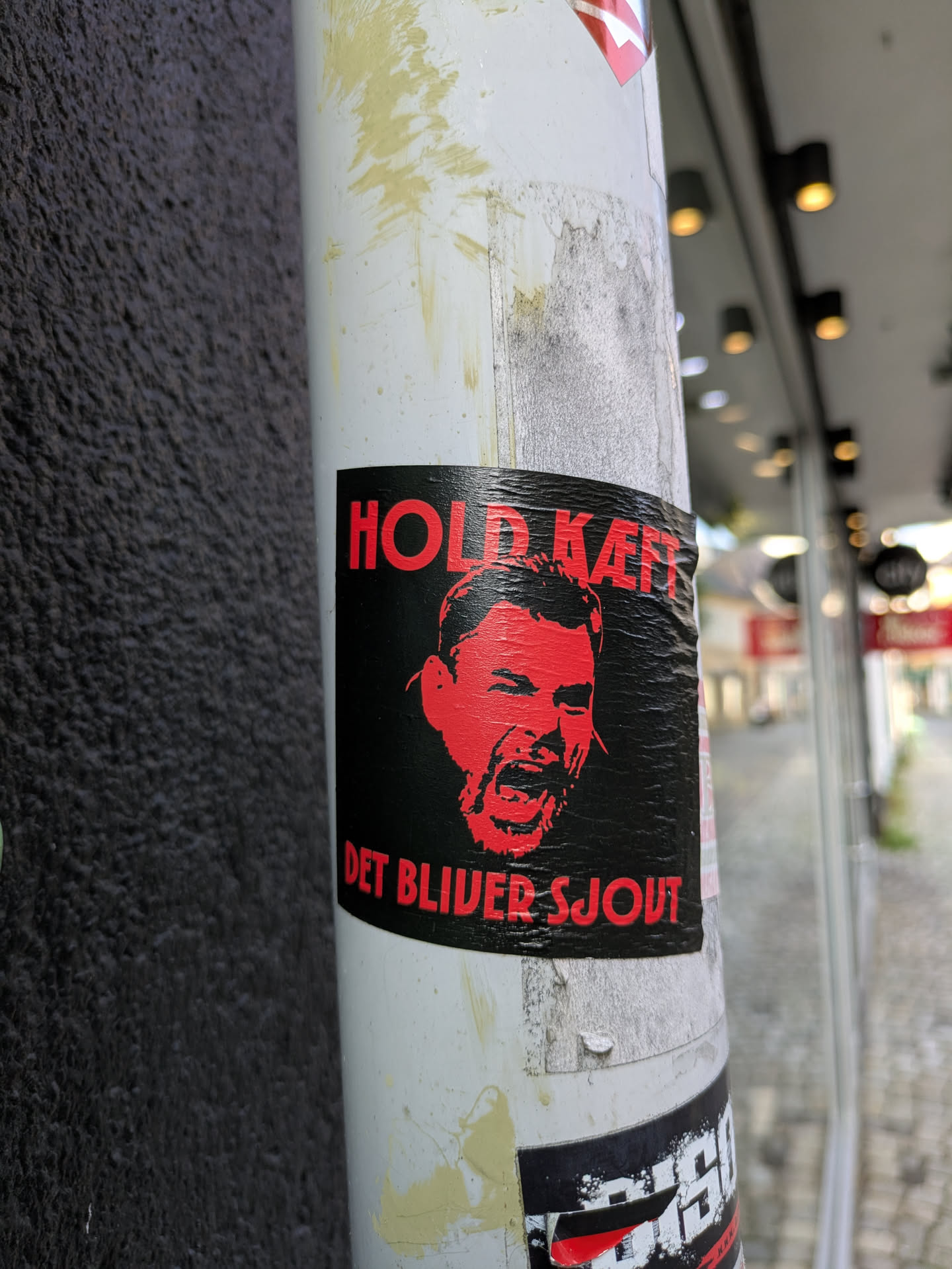 The sticker is rectangular with a stark black background, against which a vibrant red, stylized illustration of an angry or shouting face is prominently displayed. The face has an open mouth and appears to be in mid-scream or roar. Above the face, in bold red uppercase letters, is the text 'HOLD KÆFT' (Shut up). Below the face, also in red uppercase letters, is 'DET BLIVER SJOUT' (It will be fun), creating a stark, possibly sarcastic or threatening, contrast with the command. The sticker itself shows signs of wear and wrinkles, suggesting it has been exposed to the elements. It is affixed to a cylindrical white pole, which is weathered with dirt and some yellow paint splatters. In the background, a dark, textured wall is visible to the left, and a blurred street scene with building lights can be seen to the right.