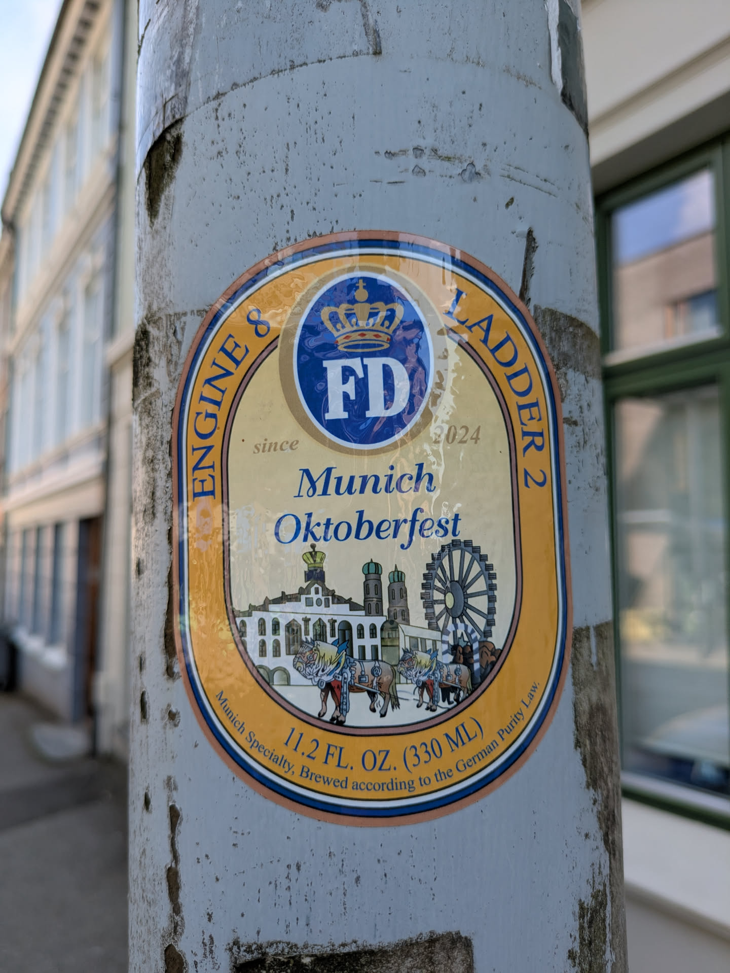 The sticker is oval-shaped with an outer orange-gold border and an inner blue border, enclosing a cream-colored background. At the top, there's a blue circle with the letters 'FD' and a crown above it, flanked by the text 'ENGINE & LADDER 2' around the curve. Below 'FD', 'since 2024' is subtly written. The central prominent text reads 'Munich Oktoberfest' in blue script. Below this, an illustration depicts a cityscape with distinctive buildings, including twin-domed towers resembling Munich's Frauenkirche, a large Ferris wheel, and horse-drawn carriages in the foreground. At the bottom, the text '11.2 FL. OZ. (330 ML)' is visible, with 'Munich Specialty, Brewed according to the German Purity Law.' curved along the lower edge. The sticker itself appears slightly worn and bubbly in places.