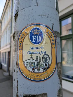 Street sticker The sticker is oval-shaped with an outer orange-gold border and an inner blue border, enclosing a cream-colored background. At the top, there's a blue circle with the letters 'FD' and a crown above it, flanked by the text 'ENGINE & LADDER 2' around the curve. Below 'FD', 'since 2024' is subtly written. The central prominent text reads 'Munich Oktoberfest' in blue script. Below this, an illustration depicts a cityscape with distinctive buildings, including twin-domed towers resembling Munich's Frauenkirche, a large Ferris wheel, and horse-drawn carriages in the foreground. At the bottom, the text '11.2 FL. OZ. (330 ML)' is visible, with 'Munich Specialty, Brewed according to the German Purity Law.' curved along the lower edge. The sticker itself appears slightly worn and bubbly in places.