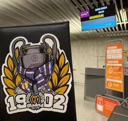 Street sticker The sticker prominently displays a stylized illustration of the UEFA Champions League trophy, rendered in metallic grey tones with black outlines and white highlights. A blue and purple striped scarf, featuring the Real Madrid crest, is wrapped around the base of the trophy. Behind the trophy, a golden laurel wreath frames the design. Below the central image, the numbers '19' and '02' are boldly presented in white with a black outline, flanking a smaller, detailed Real Madrid crest with the word 'Madrid' written beneath it. The entire sticker has a clean white border, contrasting with the black textured surface it's affixed to.