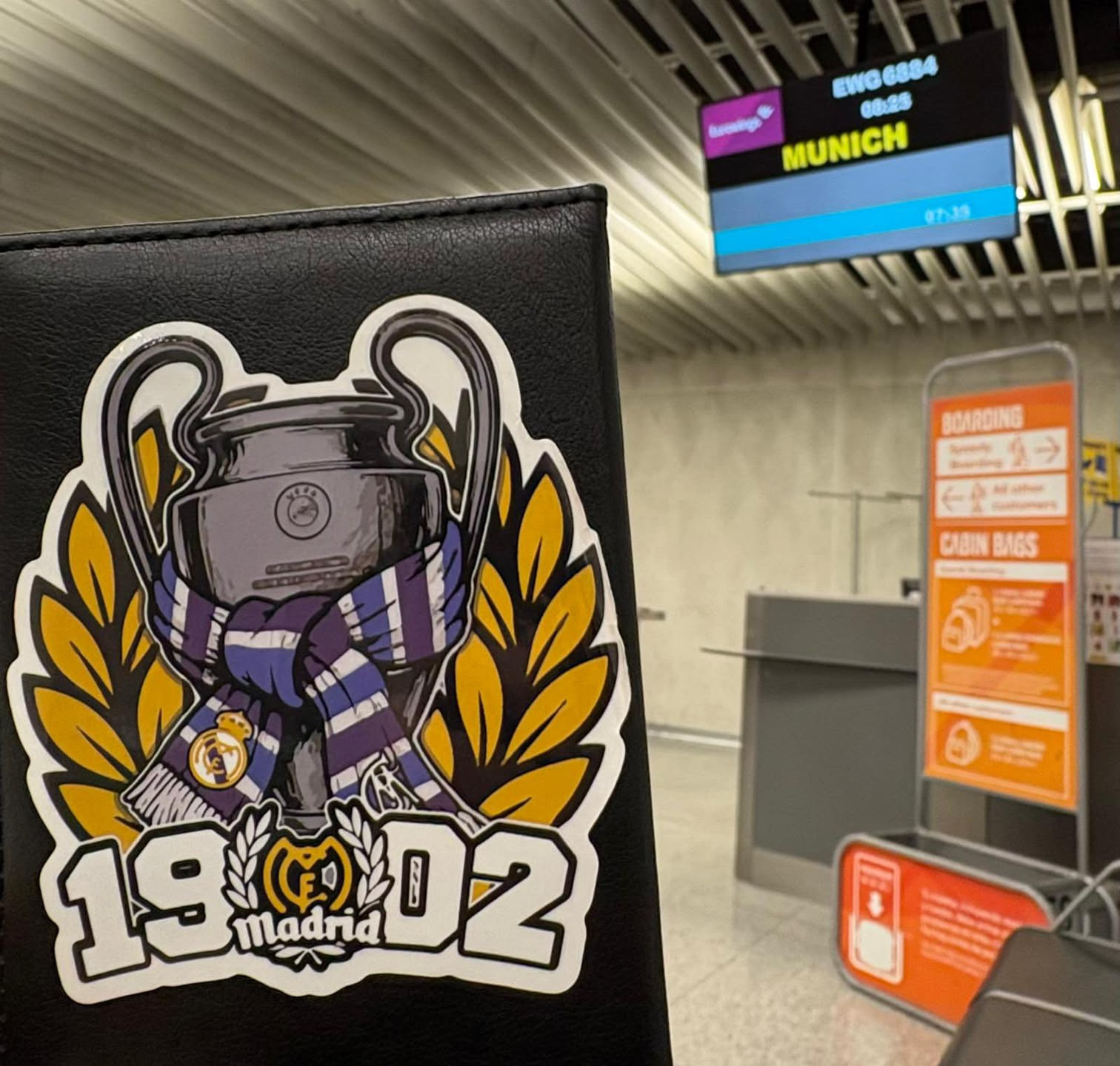 The sticker prominently displays a stylized illustration of the UEFA Champions League trophy, rendered in metallic grey tones with black outlines and white highlights. A blue and purple striped scarf, featuring the Real Madrid crest, is wrapped around the base of the trophy. Behind the trophy, a golden laurel wreath frames the design. Below the central image, the numbers '19' and '02' are boldly presented in white with a black outline, flanking a smaller, detailed Real Madrid crest with the word 'Madrid' written beneath it. The entire sticker has a clean white border, contrasting with the black textured surface it's affixed to.